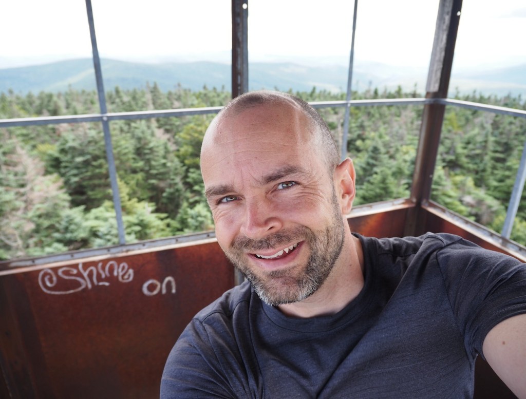 a photo of a smiling man, mostly bald with light beard, in a forest fire tower