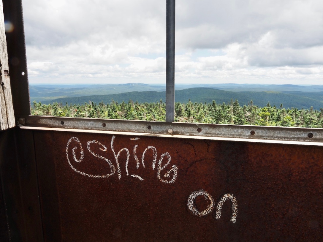 The words "shine on" written in white chalk on a rusty fire tower wall, overlooking a green forest.
