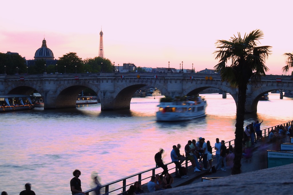 The Seine River in Paris at sunset, people on the bank and a bridge with boat going under in the foreground; Eiffel Tower in the background.